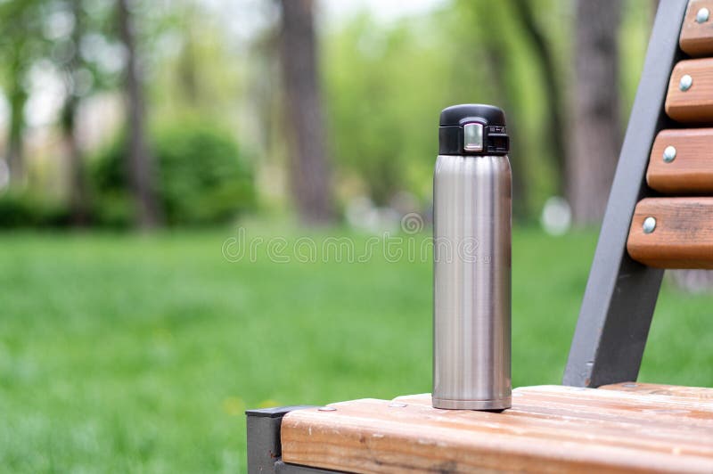 Steel thermal mug standing on a park bench. Thermos on a bench in the park. Front view. Steel thermal mug standing on a park bench. Thermos on a bench in the park. Front view.