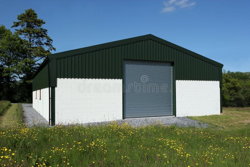 Newly constructed barn of cream painted concrete block walls with a green metal sheet roof and roller shutter doors, standing in a field of buttercups, with a blue sky and trees to the rear. Newly constructed barn of cream painted concrete block walls with a green metal sheet roof and roller shutter doors, standing in a field of buttercups, with a blue sky and trees to the rear.