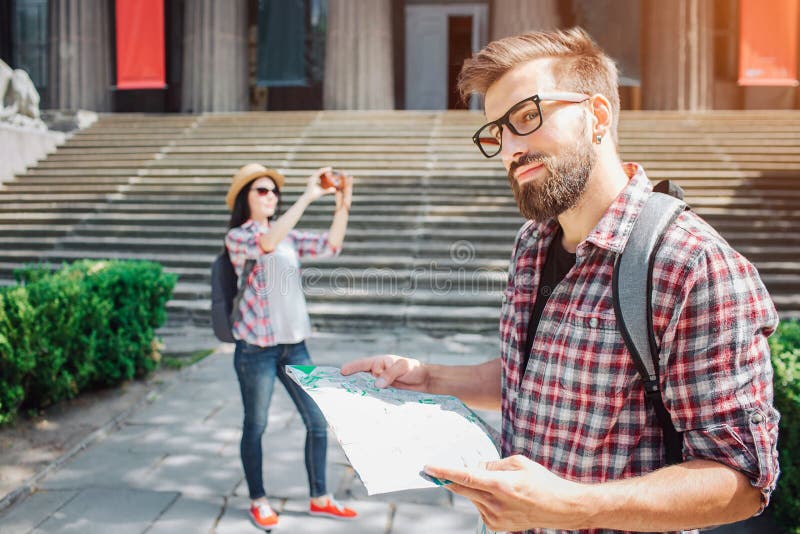 Standaußenseite des gut aussehenden Mannes und lächeln ein Stückchen Er hält Karte in den Händen Junge weibliche touristische Stä stockbild Standaußenseite des gut aussehenden Mannes und lächeln ein Stückchen Er hält Karte in den Händen Junge weibliche touristische Stä stockbild