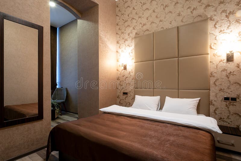 Stanza con un letto matrimoniale, il comodino, lo specchio sulla parete, la porta bianca, le pareti beige, la pavimentazione lami immagini stock libere da diritti Stanza con un letto matrimoniale, il comodino, lo specchio sulla parete, la porta bianca, le pareti beige, la pavimentazione lami immagini stock libere da diritti