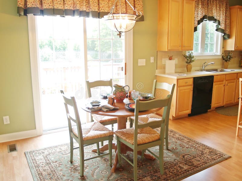 A small breakfast table in front of a bright window. The table is set for a meal, and the kitchen is in the background. A small breakfast table in front of a bright window. The table is set for a meal, and the kitchen is in the background.