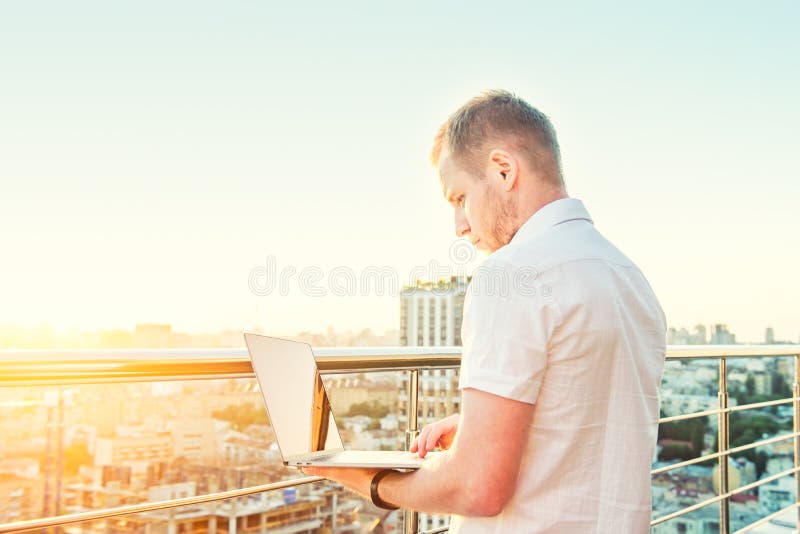 Starker junger Geschäftsmann, der an dem Laptop steht auf hohem Aufstiegsgebäudebalkon mit städtischem Stadtbild des Sonnenunterg lizenzfreie stockfotografie Starker junger Geschäftsmann, der an dem Laptop steht auf hohem Aufstiegsgebäudebalkon mit städtischem Stadtbild des Sonnenunterg lizenzfreie stockfotografie