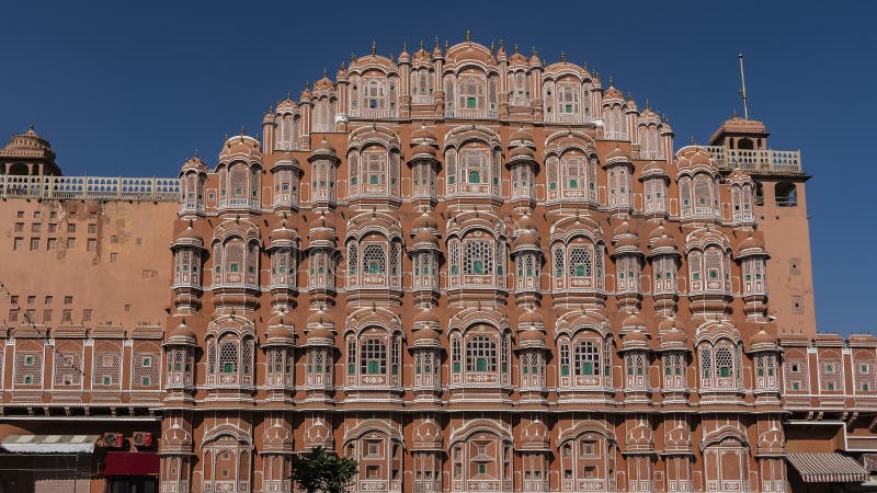 The ancient Palace of the Winds- Hawa Mahal against the blue sky. The five-storey red sandstone building in the form of the crown of Krishna. There are many windows with openwork grilles on the facade. The ancient Palace of the Winds- Hawa Mahal against the blue sky. The five-storey red sandstone building in the form of the crown of Krishna. There are many windows with openwork grilles on the facade