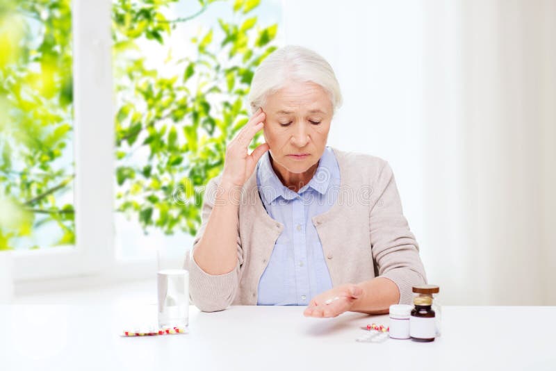 Age, medicine and healthcare concept - senior woman with pills and glass of water at home over green natural background. Age, medicine and healthcare concept - senior woman with pills and glass of water at home over green natural background