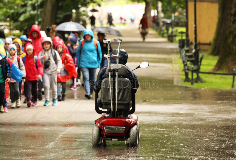 Starszy mężczyzna w machinalnym wózku inwalidzkim przechodzi alee park za przelotną grupą dzieci Współczucie i pomoc obrazy stock Starszy mężczyzna w machinalnym wózku inwalidzkim przechodzi alee park za przelotną grupą dzieci Współczucie i pomoc obrazy stock
