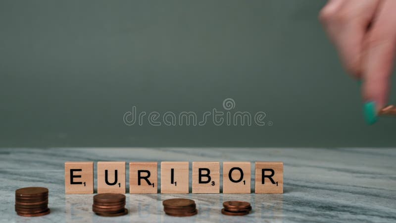Static Shot of Word EURIBOR Is Written In Wooden Letters On Grey Background. In Foreground, Woman Lays Out Piles of Euro Cents.
