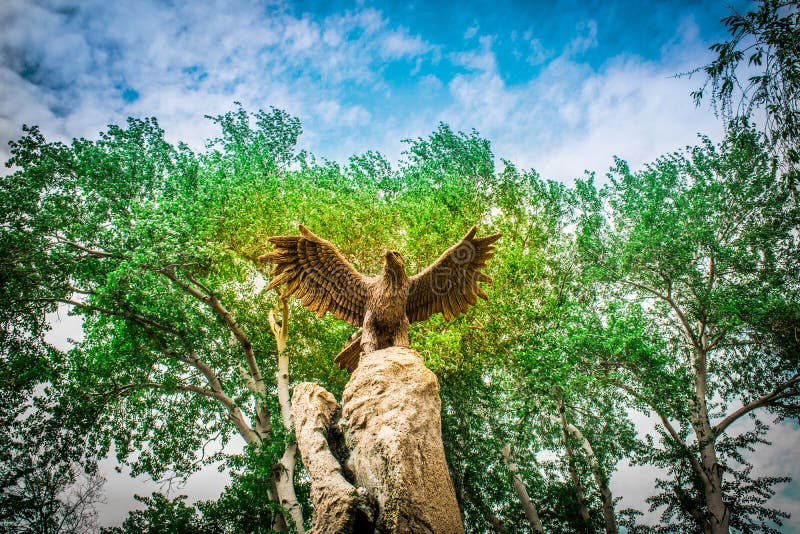 Statue of a large brown eagle with open large wings against the background of green trees and a blue cloudy sky. Bottom view. image of freedom and flight of a proud strong bird. Statue of a large brown eagle with open large wings against the background of green trees and a blue cloudy sky. Bottom view. image of freedom and flight of a proud strong bird.