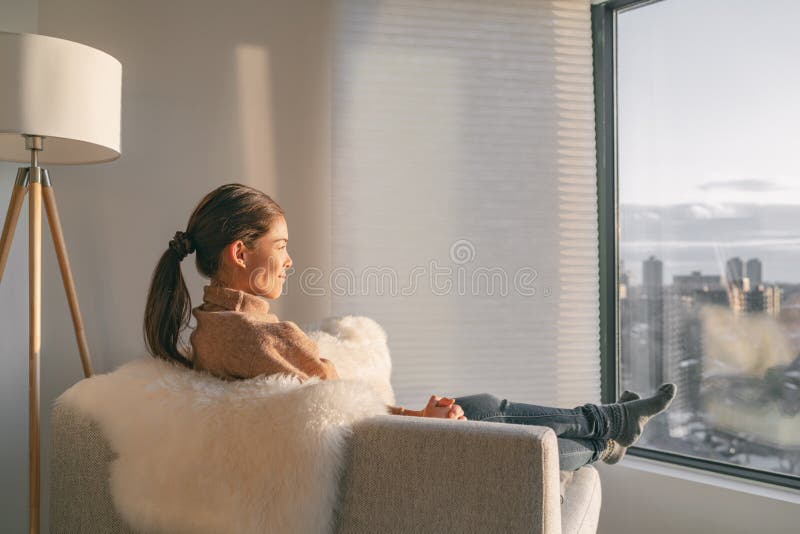 Staying home Asian woman relaxing pensive looking out the window sitting in living room sofa enjoying free time to think