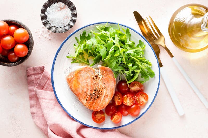 Fried pink tuna steak served with arugula and tomatoes on a plate, pink background, top view. Healthy meal rich in omega 3. Fried pink tuna steak served with arugula and tomatoes on a plate, pink background, top view. Healthy meal rich in omega 3