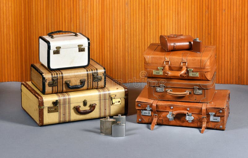 Still Life of Antique Suitcases and Flasks - Two Stacks of Various Sized Suitcases and Trunks with Collection of Metal Alcohol Drinking Flasks in Room with Gray Floor and Wood Wall Paneling. Still Life of Antique Suitcases and Flasks - Two Stacks of Various Sized Suitcases and Trunks with Collection of Metal Alcohol Drinking Flasks in Room with Gray Floor and Wood Wall Paneling