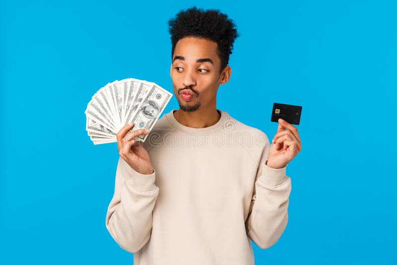 Modern lifestyle, business and finance concept. Cheerful happy african-american guy thinking how waste money, kissing cash dollars and holding credit card, winning prize, stading blue background. Modern lifestyle, business and finance concept. Cheerful happy african-american guy thinking how waste money, kissing cash dollars and holding credit card, winning prize, stading blue background.