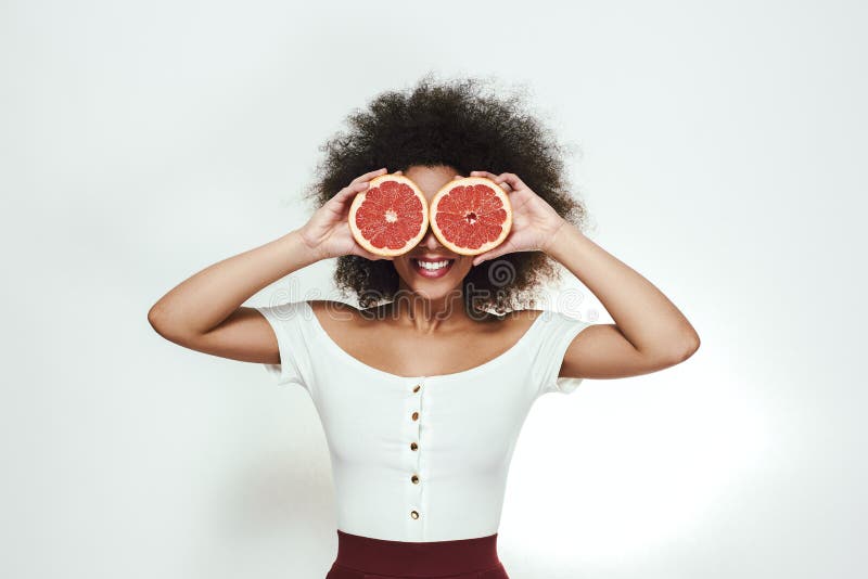 Healthy lifestyle. Playful young afro american woman holding two parts of grapefruit at her eyes and smiling while standing against grey background. ts. Healthy food concept. Healthy lifestyle. Playful young afro american woman holding two parts of grapefruit at her eyes and smiling while standing against grey background. ts. Healthy food concept