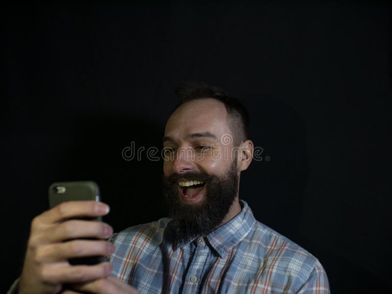 Stilfull man med ett skägg och mustaschblickar in i en mobiltelefon med en skratta framsida på en svart bakgrund royaltyfria foton Stilfull man med ett skägg och mustaschblickar in i en mobiltelefon med en skratta framsida på en svart bakgrund royaltyfria foton