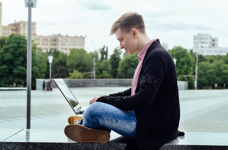 Stilfull ung man i omslaget och jeans som sitter och skriver på det fria för bärbar datordator, sidosikt Teknologi och kommunikat royaltyfri fotografi Stilfull ung man i omslaget och jeans som sitter och skriver på det fria för bärbar datordator, sidosikt Teknologi och kommunikat royaltyfri fotografi