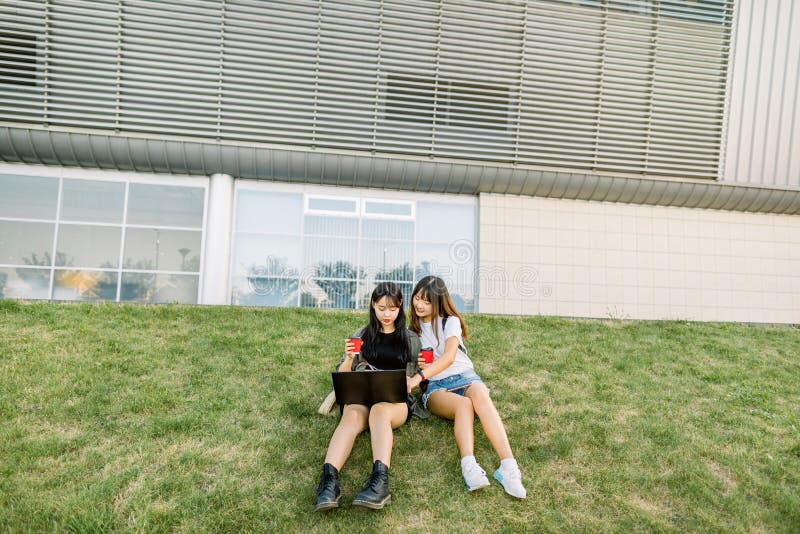 Urban lifestyle and digital technologies. Pretty young Korean girls best friends resting on the green grass, drinking coffee and working on laptop near the big urban building. Urban lifestyle and digital technologies. Pretty young Korean girls best friends resting on the green grass, drinking coffee and working on laptop near the big urban building.