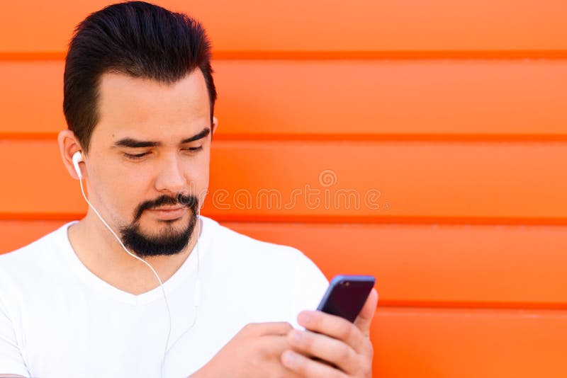 Stilig man med skägget och mustasch som lyssnar till musik eller håller ögonen på något på skärmen av hans smartphone med hörlura royaltyfri bild Stilig man med skägget och mustasch som lyssnar till musik eller håller ögonen på något på skärmen av hans smartphone med hörlura royaltyfri bild