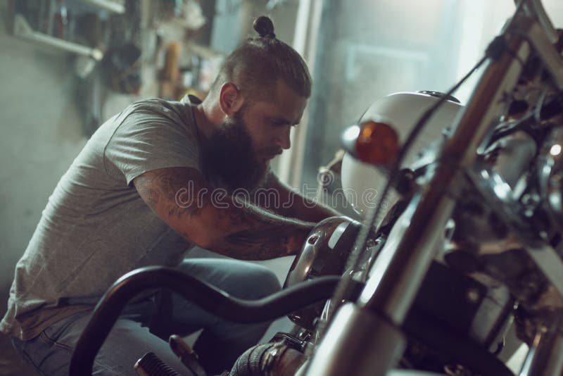 Stilig skäggig man som reparerar hans motorcykel i garaget Bärande jeans för en man och enskjorta royaltyfri bild Stilig skäggig man som reparerar hans motorcykel i garaget Bärande jeans för en man och enskjorta royaltyfri bild
