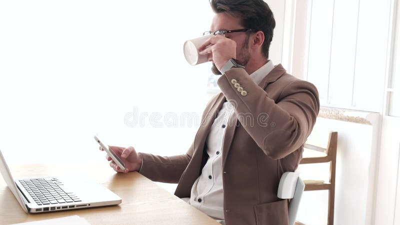 Stilig ung aff? rsman som anv? nder hans smartphone, medan dricka kaffe framme av b? rbara datorn i kontoret stock video