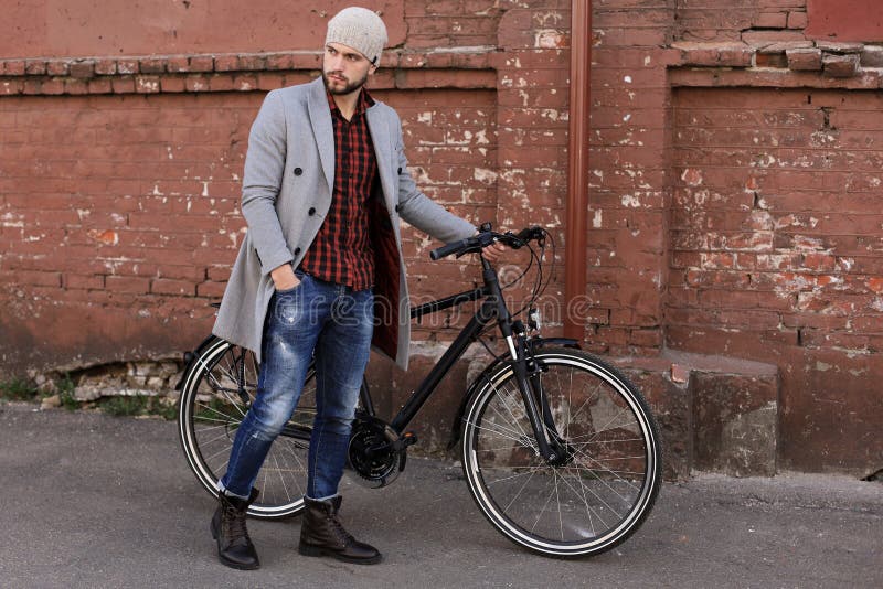 Stilig ung man i gr?tt lag och hatten som skjuter hans cykel ner en gata i staden arkivfoto Stilig ung man i gr?tt lag och hatten som skjuter hans cykel ner en gata i staden arkivfoto