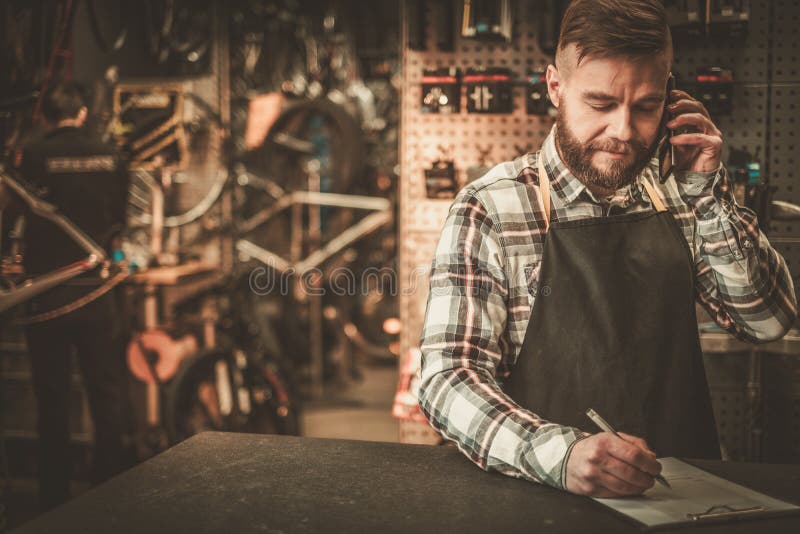 Stilvoller Fahrradmechaniker nimmt Bestellung telefonisch in seiner Werkstatt entgegen lizenzfreies stockbild Stilvoller Fahrradmechaniker nimmt Bestellung telefonisch in seiner Werkstatt entgegen lizenzfreies stockbild