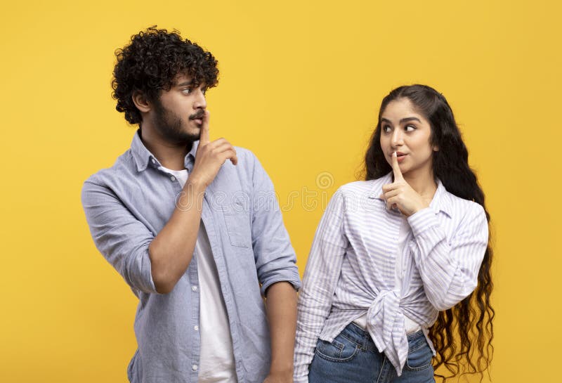 Shh, keep silence concept. Cunning indian couple gesturing hush sign, posing over yellow studio background, looking at each other, holding finger on lips. Silent gesture, secret. Shh, keep silence concept. Cunning indian couple gesturing hush sign, posing over yellow studio background, looking at each other, holding finger on lips. Silent gesture, secret