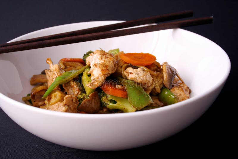 A freshly cooked chicken stir fry with fresh vegetables singapore noodles served in a white bowl with chop sticks. A freshly cooked chicken stir fry with fresh vegetables singapore noodles served in a white bowl with chop sticks