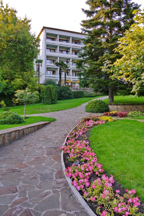 A stone path with planted flowers, tall green trees and a lawn leads to a white five-story building with balconies.