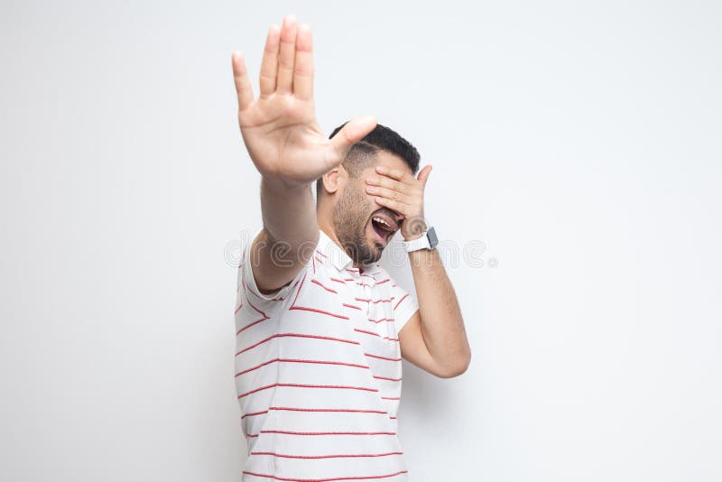 Stop, i don`t want to look at this. Portrait of confused bearded young man in striped t-shirt standing, covering his eyes and stock image Stop, i don`t want to look at this. Portrait of confused bearded young man in striped t-shirt standing, covering his eyes and stock image