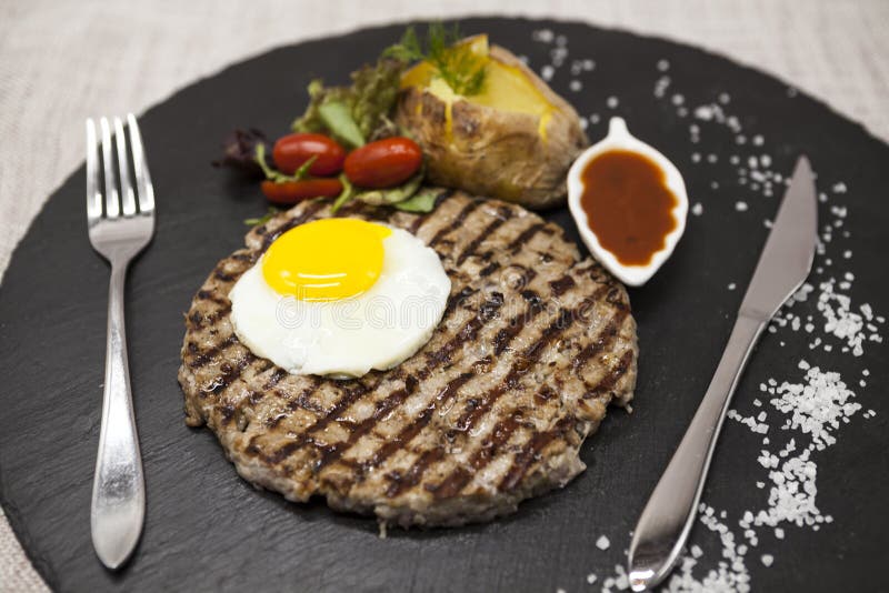 Stor, saftig läcker stek med ett stekt ägg och bakade potatisar på en stenplatta arkivfoto Stor, saftig läcker stek med ett stekt ägg och bakade potatisar på en stenplatta arkivfoto