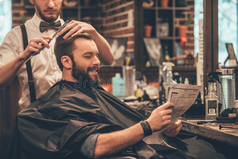 Cheerful young bearded men getting haircut by hairdresser and reading newspaper while sitting in chair at barbershop. Cheerful young bearded men getting haircut by hairdresser and reading newspaper while sitting in chair at barbershop