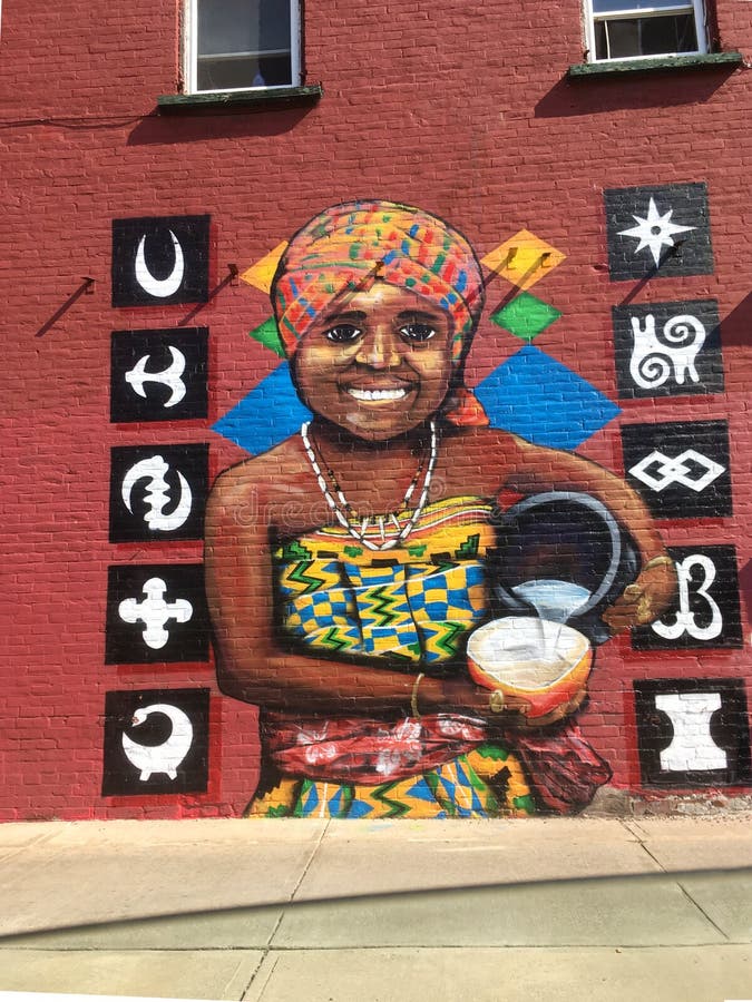 An image of a large painted mural on the side of an African Grocer in the old north end of Burlington, Vermont depicting an African woman in traditional dress pouring water into a gourd with many of the largest African Tribal symbols such as the Ashanti. An image of a large painted mural on the side of an African Grocer in the old north end of Burlington, Vermont depicting an African woman in traditional dress pouring water into a gourd with many of the largest African Tribal symbols such as the Ashanti.