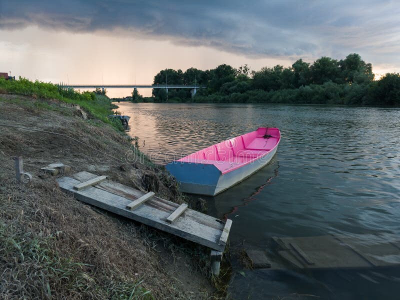 Storm arcus shaft and cumulonimbus cloud with heavy rain or summer shower, severe weather and sun glow behind rain. Landscape with Sava river with moored boat next to wooden dock and bridge.