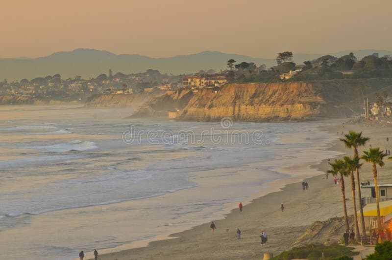 The beach at Del Mar, southern California, near San Diego, on the west coast of the United States, in the soft rays of the sun sunset, with the sun turning the buildings and cliffs orange and with people walking the sand and standing outside buildings to see the sun hit the ocean. The beach at Del Mar, southern California, near San Diego, on the west coast of the United States, in the soft rays of the sun sunset, with the sun turning the buildings and cliffs orange and with people walking the sand and standing outside buildings to see the sun hit the ocean.