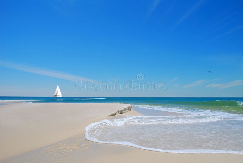 Pretty beach sand and dune with gentle waves with sailboat in distance under blue sky. Pretty beach sand and dune with gentle waves with sailboat in distance under blue sky