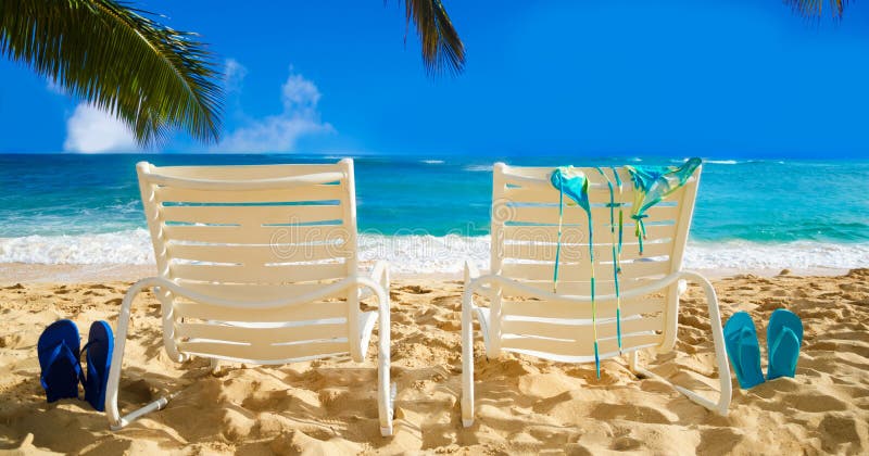 Two white beach chairs under palm leaves by the ocean, with bikini and flip flops. Two white beach chairs under palm leaves by the ocean, with bikini and flip flops.