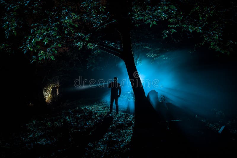 Strange light in a dark forest at night. Silhouette of person standing in the dark forest with light. Horror halloween concept.