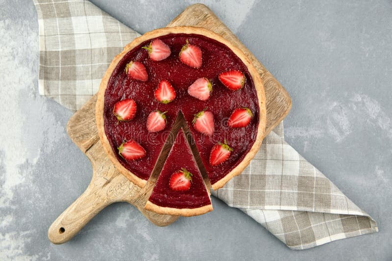 Strawberry pie with fresh berries halves on grey stone background. Sweet summer desser on wooden cutting board, top view. Piece of pie, napkin. flat lay