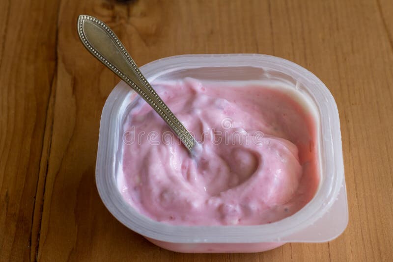 Strawberry yogurt in a plastic pot with silver spoon on rustic wooden table background - side view