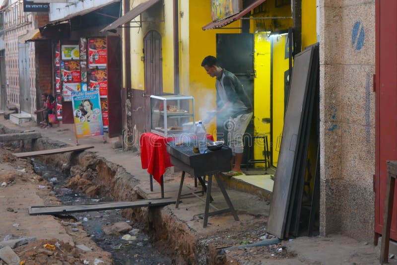 Antananarivo, Madagascar, Africa - 01/11/20: An african man cooks with a smoking grill over an open sewage system. Big hole in the street dirty water flowing. Malagasy restaurant, guy sells food on the sidewalk in an open-air market. Antananarivo, Madagascar, Africa - 01/11/20: An african man cooks with a smoking grill over an open sewage system. Big hole in the street dirty water flowing. Malagasy restaurant, guy sells food on the sidewalk in an open-air market.