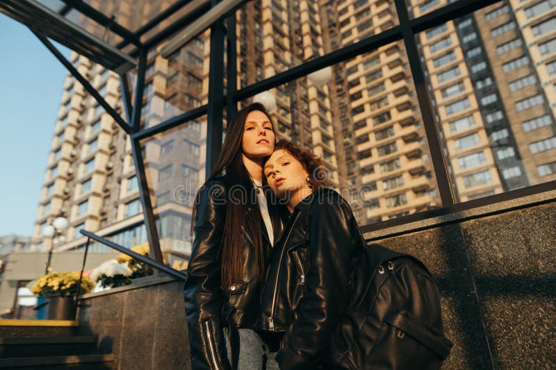 Fashion Portrait Of Two Stylish Girls Standing Against A Black Wall Hugging And Posing At The