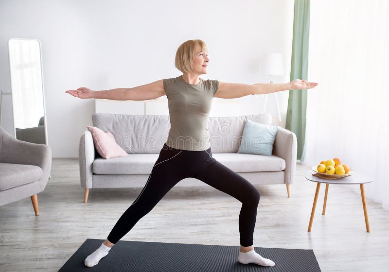 Strength yoga exercises. Fit mature lady standing in warrior asana, exercising on sports mat at home. Fit senior woman doing domestic workout, practicing pilates during coronavirus lockdown