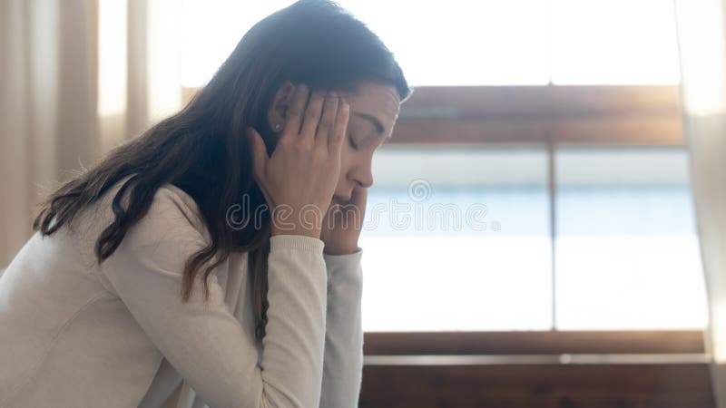 Awful day! Profile view of stressed depressed frustrated teen girl sitting at home alone with closed eyes, leaning forward hugging head having problem with parents, feeling bad or headache. Copy space. Awful day! Profile view of stressed depressed frustrated teen girl sitting at home alone with closed eyes, leaning forward hugging head having problem with parents, feeling bad or headache. Copy space