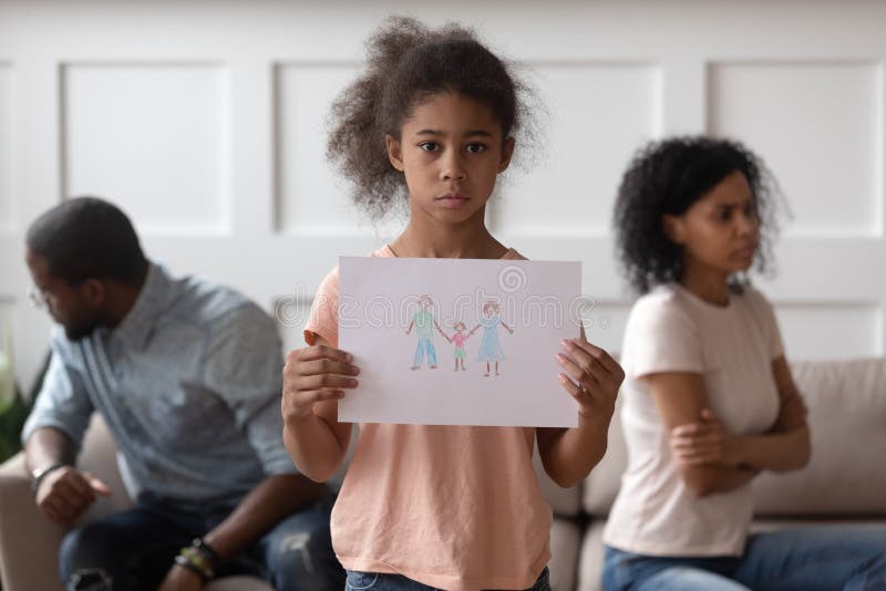 Little sad african american kid girl holding hand drawn picture with happy family, suffering from parental divorce, conflict, shared custody, mother and father separation, psychological trauma. Little sad african american kid girl holding hand drawn picture with happy family, suffering from parental divorce, conflict, shared custody, mother and father separation, psychological trauma.