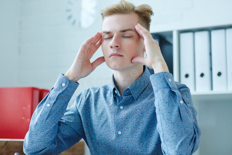 Frustrated young caucasian man massaging temple and keeping eyes closed while sitting at his working place. royalty free stock photography Frustrated young caucasian man massaging temple and keeping eyes closed while sitting at his working place. royalty free stock photography
