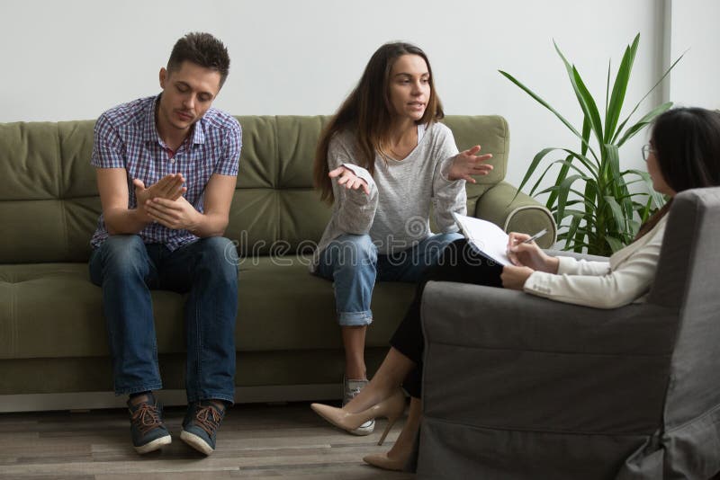 Stressed wife discussing relationship problems with female psychologist, explaining thoughts, husband indifferent, couch listening attentively. Couple getting help to save marriage, avoid break up. Stressed wife discussing relationship problems with female psychologist, explaining thoughts, husband indifferent, couch listening attentively. Couple getting help to save marriage, avoid break up