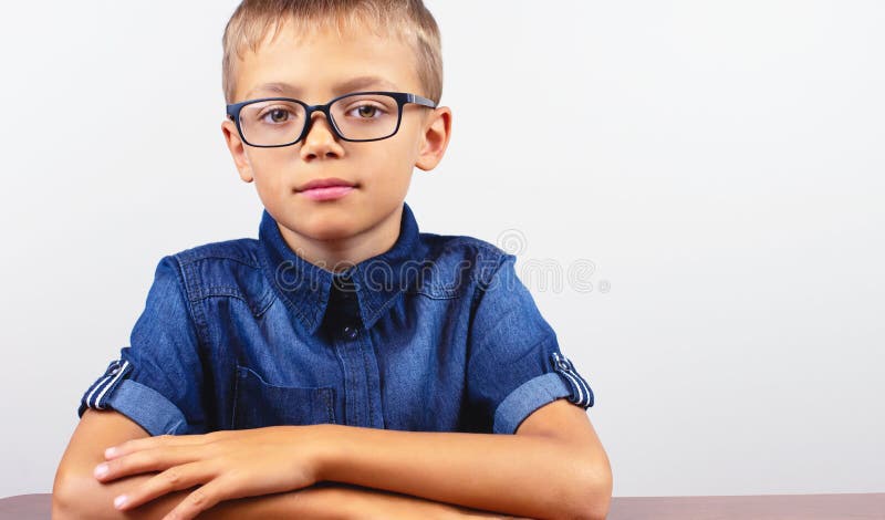 Striscione Scuolboy in camicia blu seduta al tavolo Ragazzo con occhiali su fondo bianco Concetto di nuovo a scuola fotografie stock Striscione Scuolboy in camicia blu seduta al tavolo Ragazzo con occhiali su fondo bianco Concetto di nuovo a scuola fotografie stock