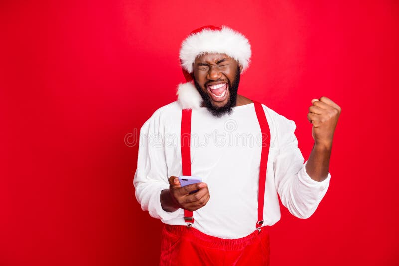 Portrait of crazy african christmas father raising fists screaming yes, got noel news like holding cell phone wearing hat white pullover isolated over red background. Portrait of crazy african christmas father raising fists screaming yes, got noel news like holding cell phone wearing hat white pullover isolated over red background