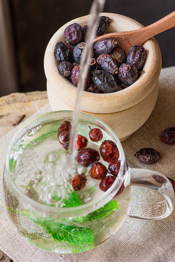 Strömendes Heißwasser in die Glasschale, die Rose Hip Tea mit frische Minzen-Beeren hölzerne Schüssel-in der Leinentuch-Ganzheits lizenzfreie stockfotografie Strömendes Heißwasser in die Glasschale, die Rose Hip Tea mit frische Minzen-Beeren hölzerne Schüssel-in der Leinentuch-Ganzheits lizenzfreie stockfotografie