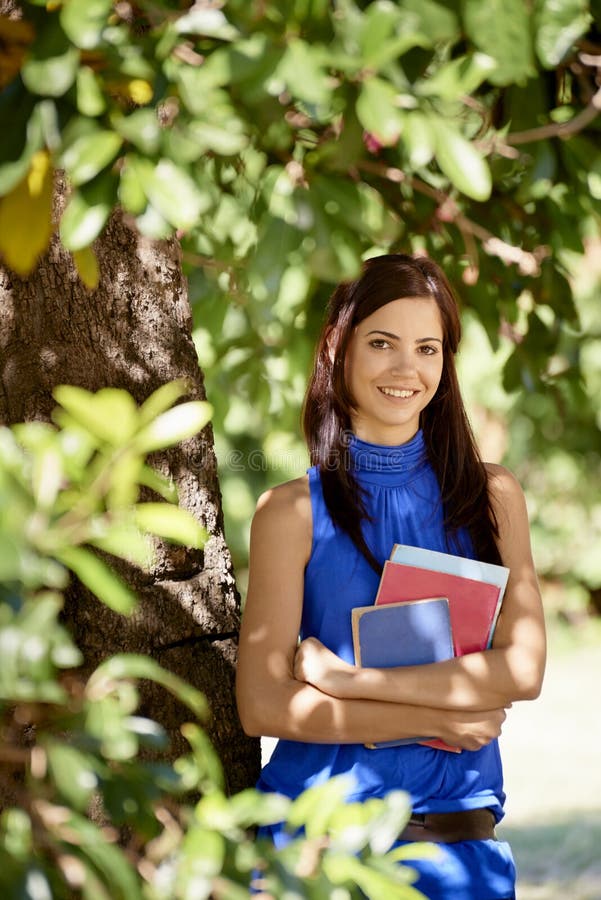 Poradie študentov portrét v škole, šťastná mladá žena s úsmevom, s college učebnice v parku oprel sa o strom.