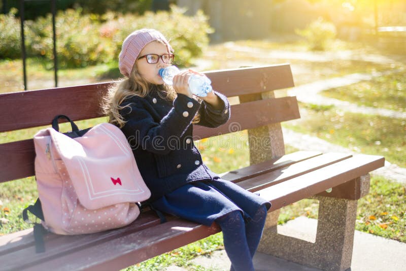 Studente della scuola elementare della bambina che si siede sul banco con lo zaino, acqua potabile dalla bottiglia immagine stock Studente della scuola elementare della bambina che si siede sul banco con lo zaino, acqua potabile dalla bottiglia immagine stock